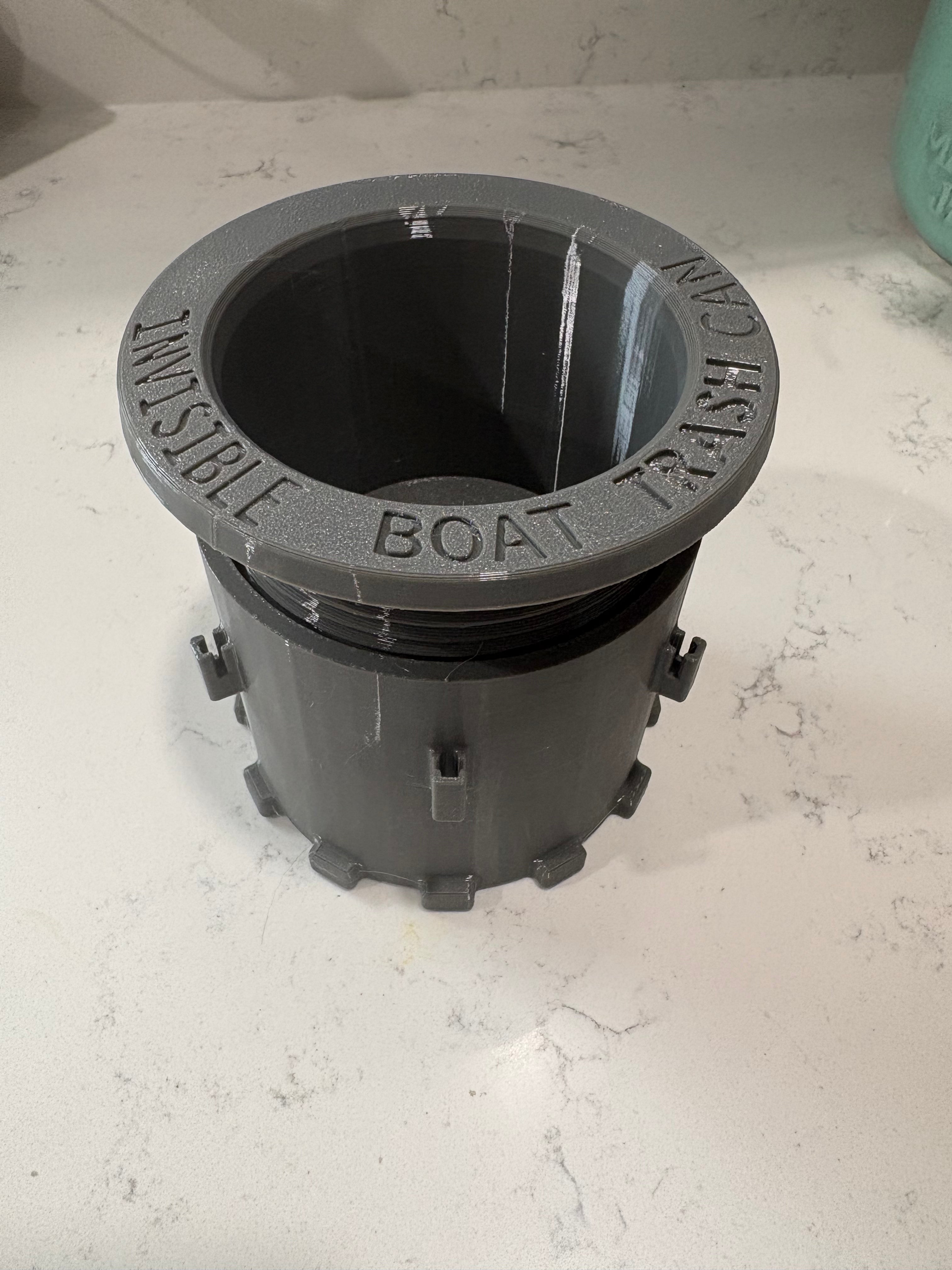 A dark gray plastic boat trash can opener assembly, featuring the words 'INVISIBLE BOAT TRASH' molded into the top rim, resting on a white marble countertop.